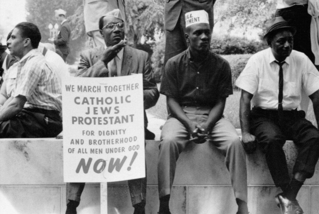 Some participants in the civil rights march sitting on a wall resting, one holds a placard which reads, "We march together, Catholics, Jews, Protestant, for dignity and brotherhood of all men under God, Now!"